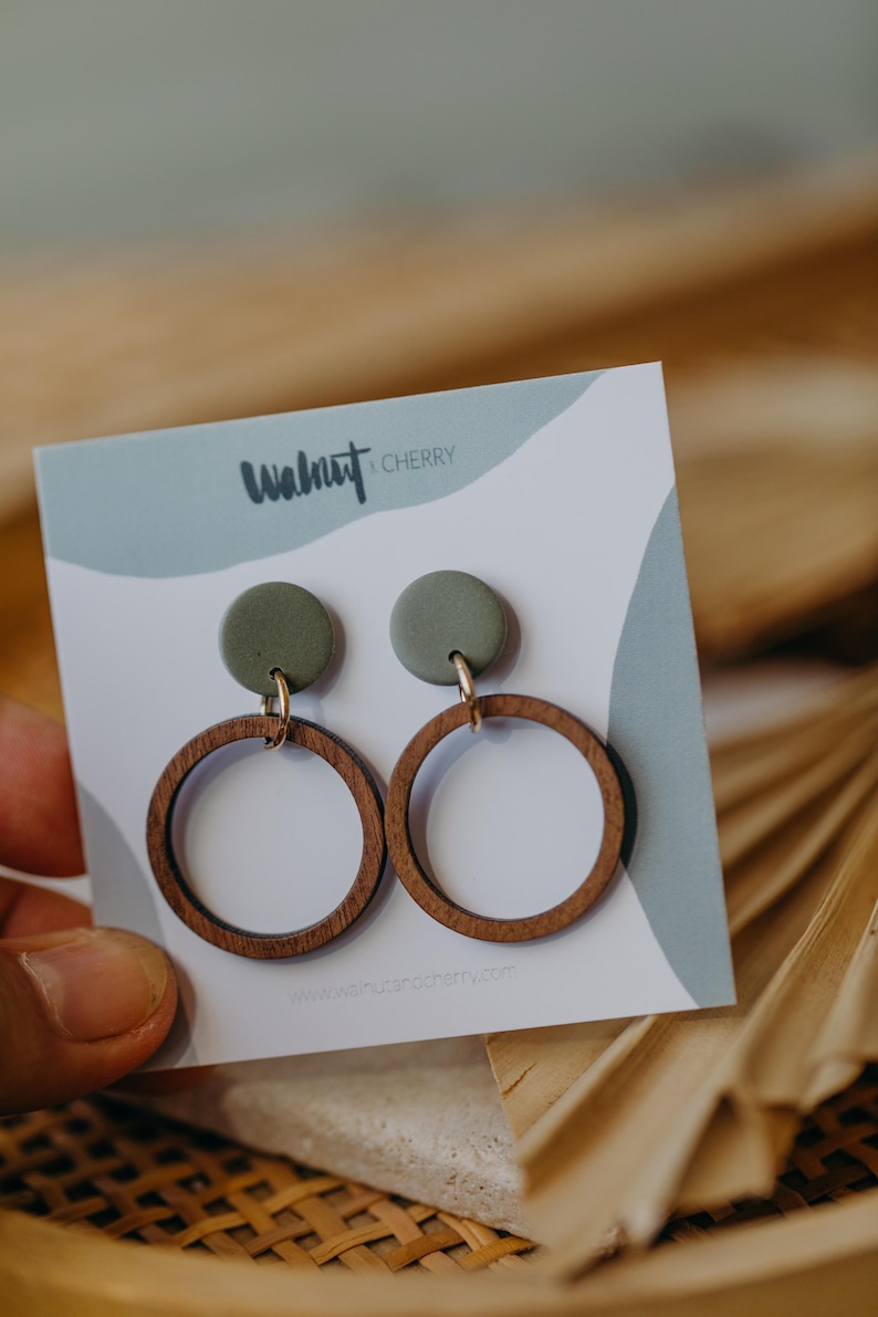 May include: A pair of green and brown wooden earrings with a round shape. The earrings are on a white card with the text "Walnut & Cherry" and a website address.