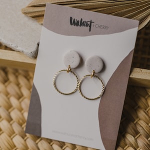 May include: A pair of gold hoop earrings with a speckled white and gray clay charm hanging from each hoop. The earrings are displayed on a white card with the text "Walnut + Cherry" printed on it.
