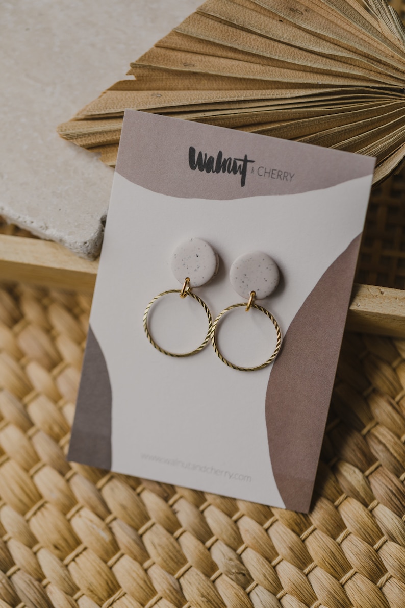 May include: A pair of gold hoop earrings with a speckled white and gray clay charm hanging from each hoop. The earrings are displayed on a white card with the text "Walnut + Cherry" printed on it.