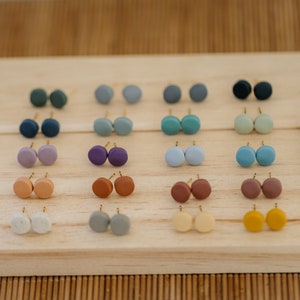 May include: A collection of 20 pairs of small, round, colorful earrings displayed on a wooden surface. The earrings are made of clay and have a matte finish.