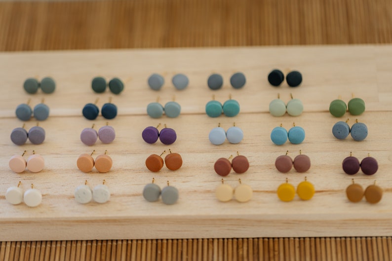 May include: A collection of 20 pairs of small, round, colorful earrings displayed on a wooden surface. The earrings are made of clay and have a matte finish.