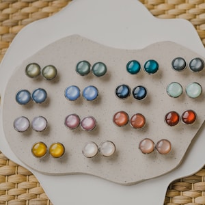 Op de afbeelding: Een display met 12 paar iriserende ronde oorbel studs in verschillende kleuren, waaronder blauw, groen, rood, roze en wit. De oorbel studs zijn in paren gerangschikt op een wit, getextureerd oppervlak.