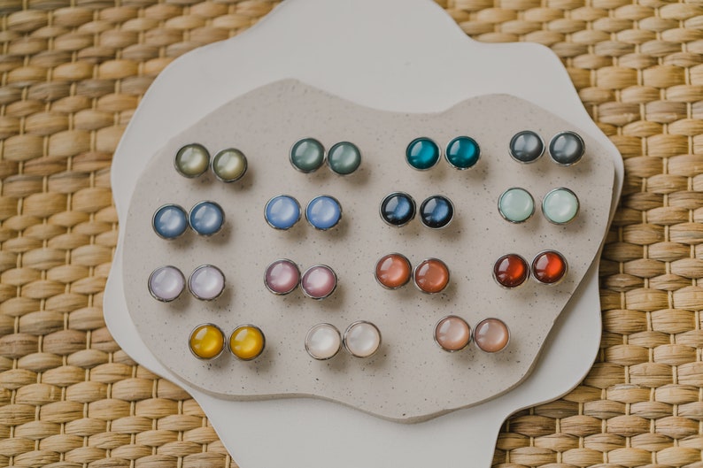 Op de afbeelding: Een display met 12 paar iriserende ronde oorbel studs in verschillende kleuren, waaronder blauw, groen, rood, roze en wit. De oorbel studs zijn in paren gerangschikt op een wit, getextureerd oppervlak.