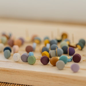 May include: A collection of colorful, round, polymer clay earrings. The earrings are scattered on a wooden surface. The earrings are various shades of blue, green, purple, brown, and beige.