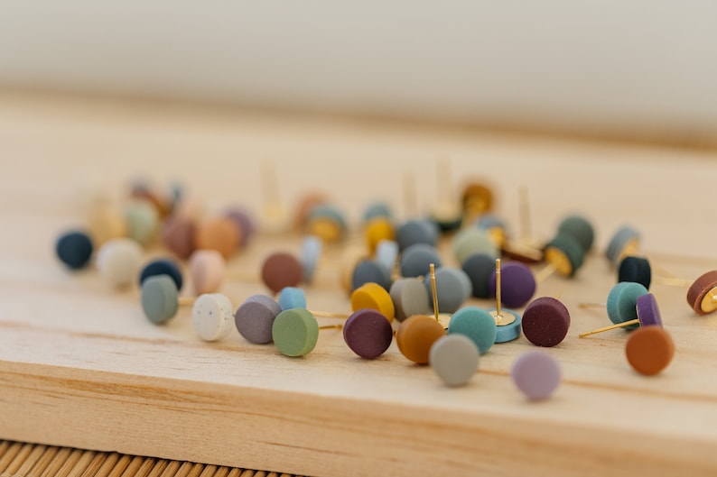 May include: A collection of colorful, round, polymer clay earrings. The earrings are scattered on a wooden surface. The earrings are various shades of blue, green, purple, brown, and beige.