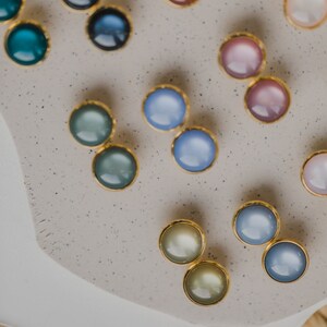 May include: Assortment of colorful round earrings with gold settings. The earrings are displayed on a light gray surface with a speckled texture.