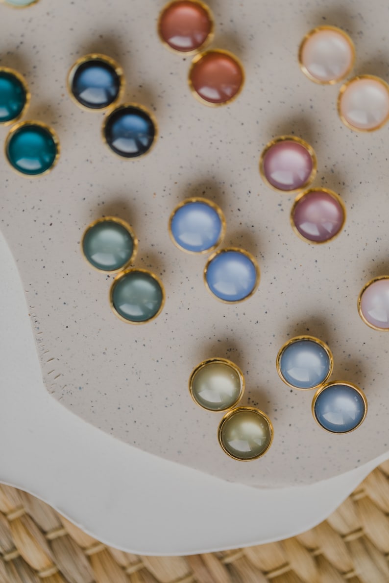 May include: Assortment of colorful round earrings with gold settings. The earrings are displayed on a light gray surface with a speckled texture.