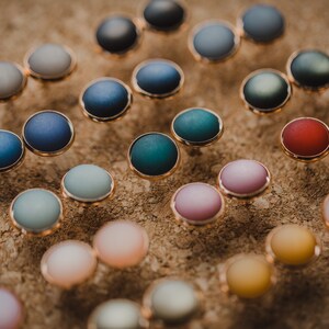 Pu&ograve; includere: Assortimento di orecchini rotondi colorati con montature dorate. Gli orecchini sono esposti su una superficie marrone.
