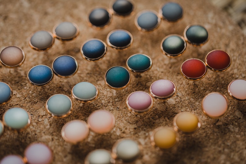 Pu&ograve; includere: Assortimento di orecchini rotondi colorati con montature dorate. Gli orecchini sono esposti su una superficie marrone.