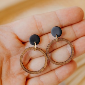 May include: A pair of wooden hoop earrings with a dark brown finish. Each earring features a small, round black accent at the top, connected to the hoop with a gold-colored finding. The earrings are displayed against a soft, neutral background.