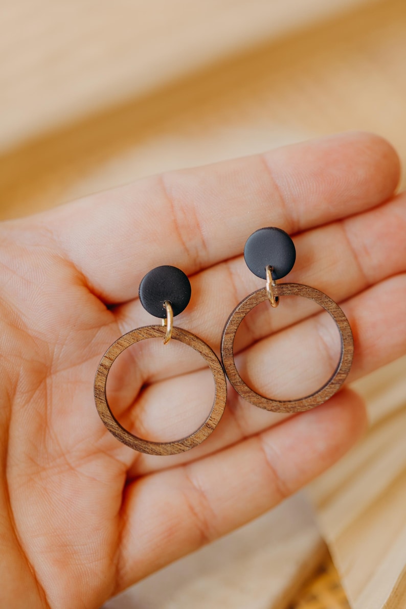 May include: A pair of wooden hoop earrings with a dark brown finish. Each earring features a small, round black accent at the top, connected to the hoop with a gold-colored finding. The earrings are displayed against a soft, neutral background.