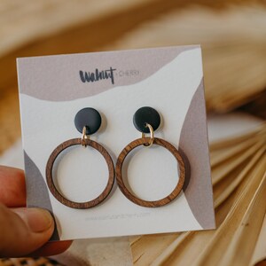 May include: A pair of wooden hoop earrings with black circular tops and gold-colored hooks. The earrings are displayed on a card with the text "Walnut Cherry" and a website address. The background is a neutral color with a palm leaf.