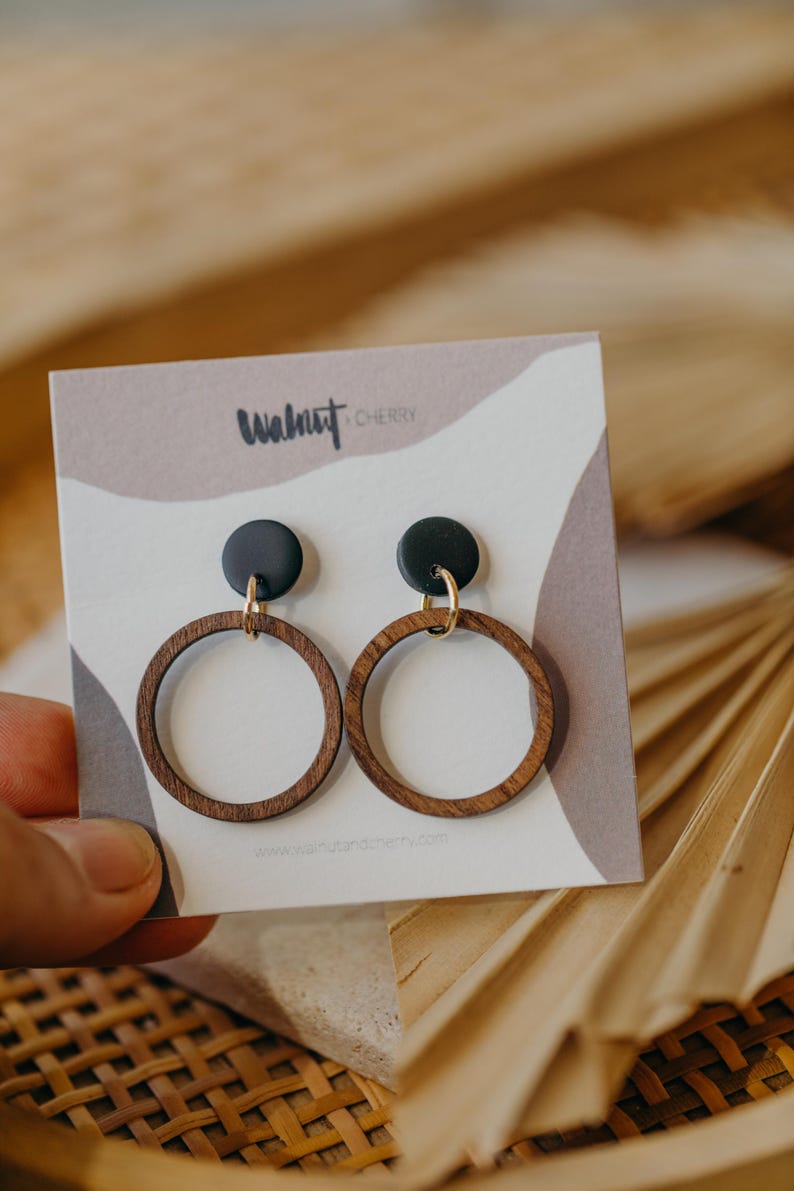 May include: A pair of wooden hoop earrings with black circular tops and gold-colored hooks. The earrings are displayed on a card with the text "Walnut Cherry" and a website address. The background is a neutral color with a palm leaf.