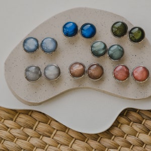 May include: A set of 10 pairs of round, iridescent earrings in various colors, including blue, green, gray, brown, and pink. The earrings are displayed on a white, textured surface.
