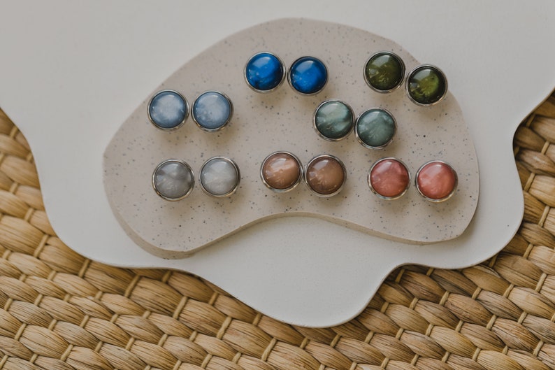 May include: A set of 10 pairs of round, iridescent earrings in various colors, including blue, green, gray, brown, and pink. The earrings are displayed on a white, textured surface.