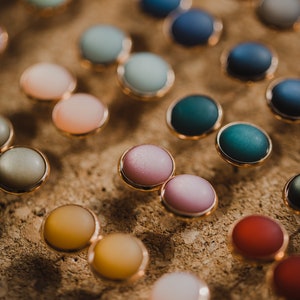 Pu&ograve; includere: Assortimento di bottoni rotondi colorati con bordo dorato. I bottoni sono disposti in file su una superficie marrone testurizzata.