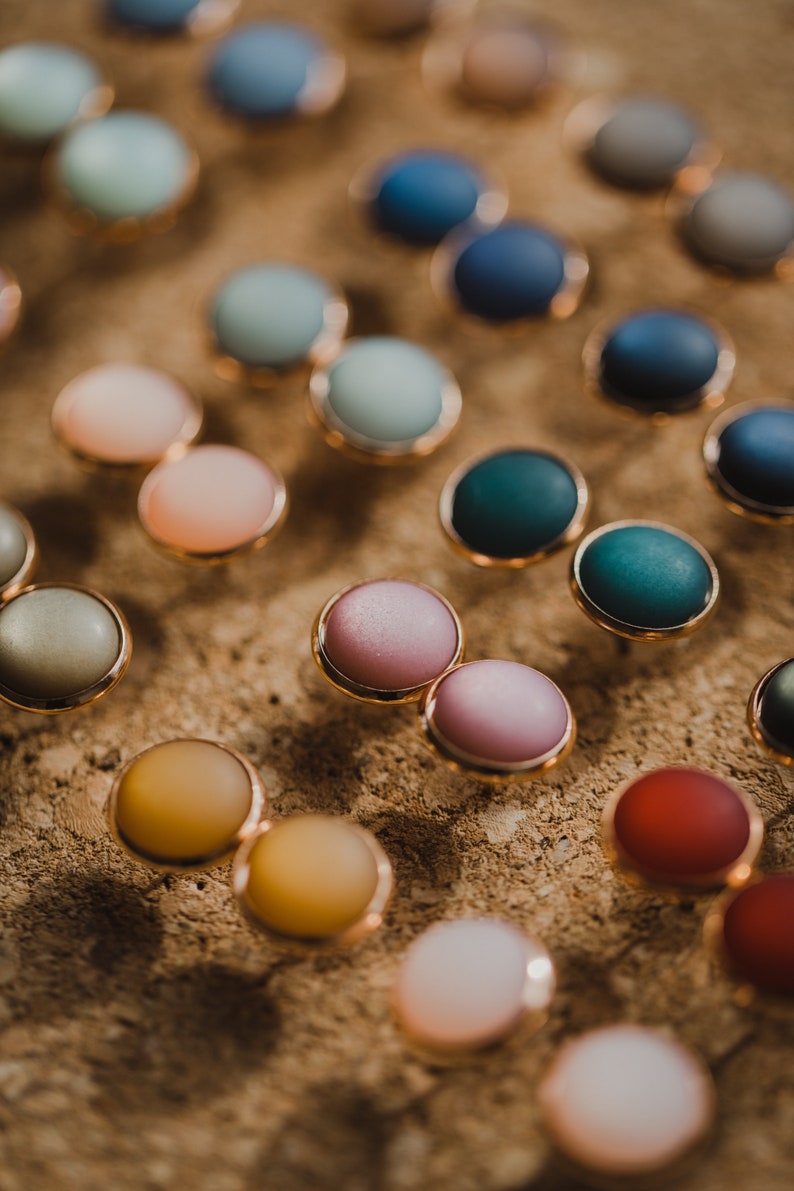 Pu&ograve; includere: Assortimento di bottoni rotondi colorati con bordo dorato. I bottoni sono disposti in file su una superficie marrone testurizzata.