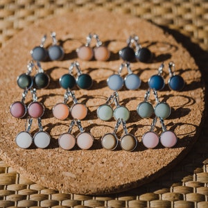 May include: A collection of 12 pairs of earrings with different colored glass cabochons. The earrings are displayed on a cork board.