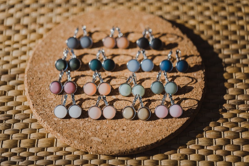 May include: A collection of 12 pairs of earrings with different colored glass cabochons. The earrings are displayed on a cork board.