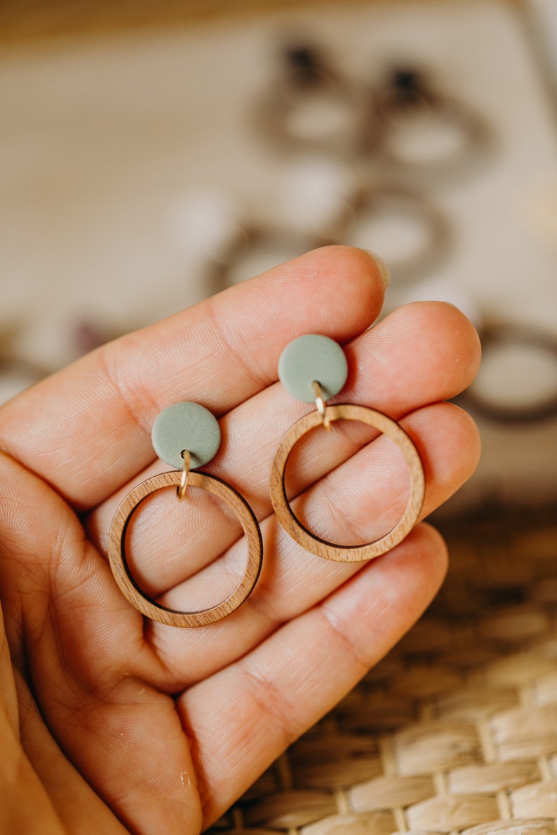 May include: A pair of wooden hoop earrings with a light green clay accent. The earrings are held in a hand.