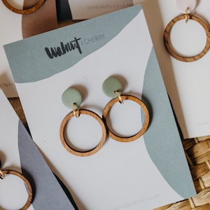 May include: A pair of wooden earrings with green clay accents. The earrings are on a white card with the text "Walnut + Cherry" and a website address.