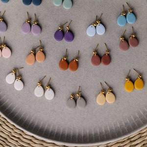 May include: A collection of 18 pairs of teardrop-shaped earrings in various colors, including pink, purple, blue, brown, gray, white, and yellow. The earrings are made of clay and have gold-colored posts.