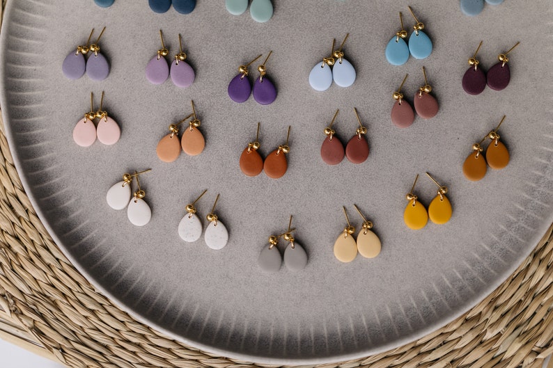 May include: A collection of 18 pairs of teardrop-shaped earrings in various colors, including pink, purple, blue, brown, gray, white, and yellow. The earrings are made of clay and have gold-colored posts.