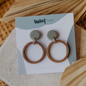 May include: A pair of green and brown wooden hoop earrings with a small green circle on top of each hoop. The earrings are displayed on a white card with the text "Walnut & Cherry" printed on it.