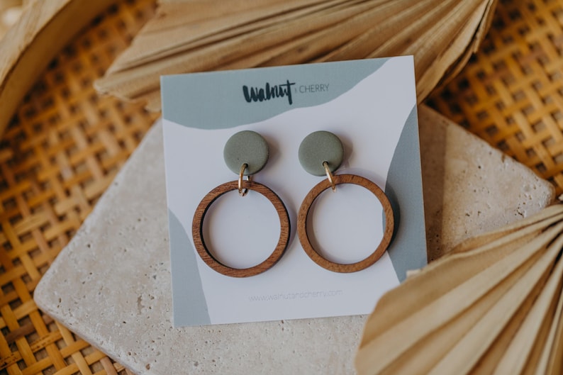 May include: A pair of green and brown wooden hoop earrings with a small green circle on top of each hoop. The earrings are displayed on a white card with the text "Walnut & Cherry" printed on it.