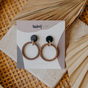 May include: A pair of wooden hoop earrings with black studs and gold-colored accents. The earrings are displayed on a card with the text "Walnut & Cherry" and a neutral color palette. The earrings are on a stone surface.