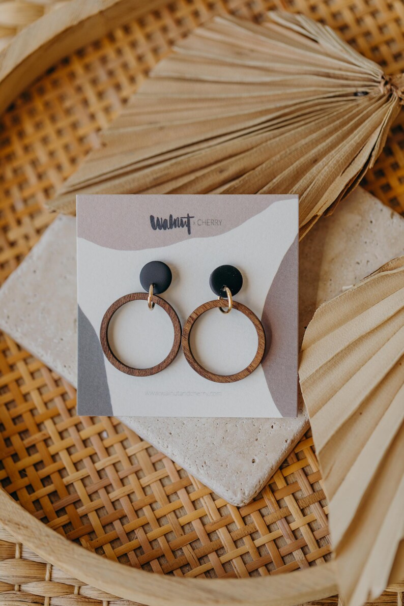 May include: A pair of wooden hoop earrings with black studs and gold-colored accents. The earrings are displayed on a card with the text "Walnut & Cherry" and a neutral color palette. The earrings are on a stone surface.