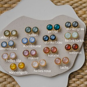May include: A display of 16 pairs of small round earrings in various colors, including pink, blue, green, yellow, and red. Each pair is labeled with a German word describing the color.