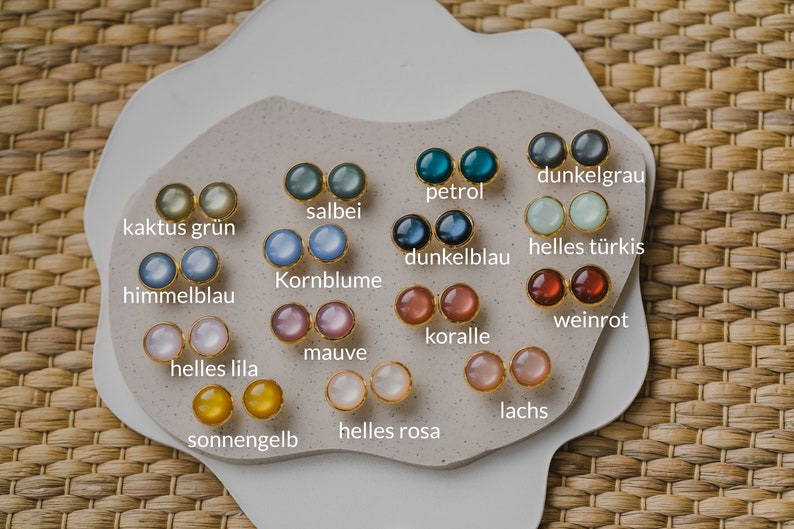 May include: A display of 16 pairs of small round earrings in various colors, including pink, blue, green, yellow, and red. Each pair is labeled with a German word describing the color.