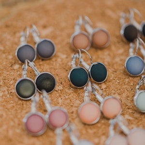 May include: Assortment of colorful round earrings with silver hooks. The earrings are displayed on a cork surface.