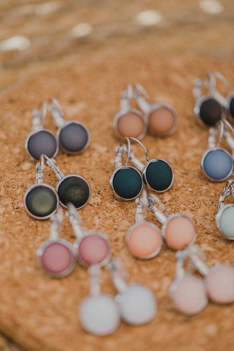 May include: Assortment of colorful round earrings with silver hooks. The earrings are displayed on a cork surface.