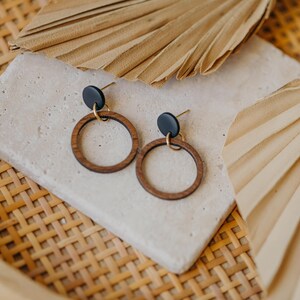 May include: A pair of wooden hoop earrings with black studs and gold-colored accents. The earrings are displayed on a light-colored, textured surface, with dried palm leaves and a woven basket in the background.