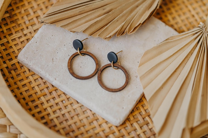 May include: A pair of wooden hoop earrings with black studs and gold-colored accents. The earrings are displayed on a light-colored, textured surface, with dried palm leaves and a woven basket in the background.