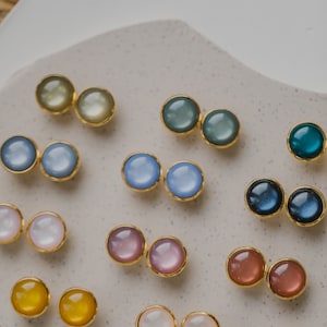 May include: Assortment of colorful round earrings with gold settings. The earrings are arranged in pairs on a white surface.