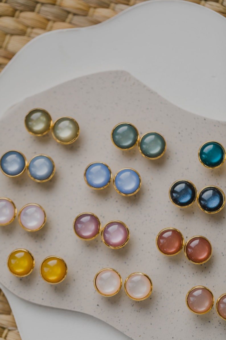 May include: Assortment of colorful round earrings with gold settings. The earrings are arranged in pairs on a white surface.