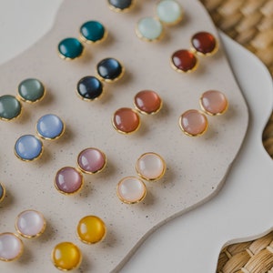May include: A display of small, round, colorful earrings with gold settings. The earrings are arranged in pairs on a white and beige surface.