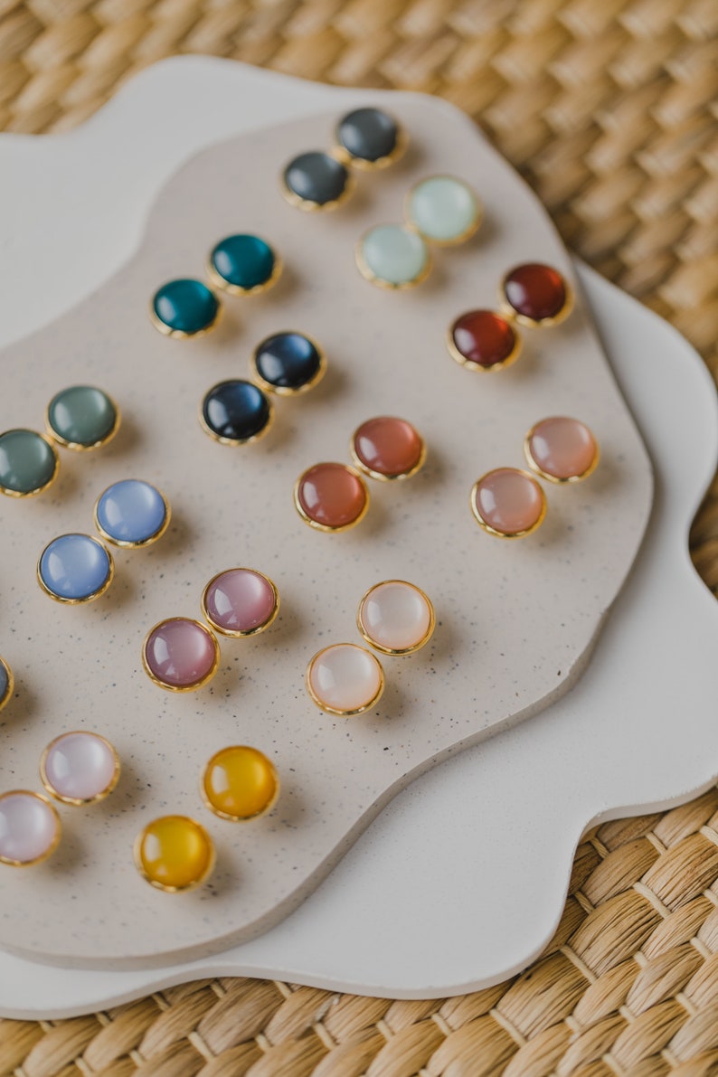May include: A display of small, round, colorful earrings with gold settings. The earrings are arranged in pairs on a white and beige surface.