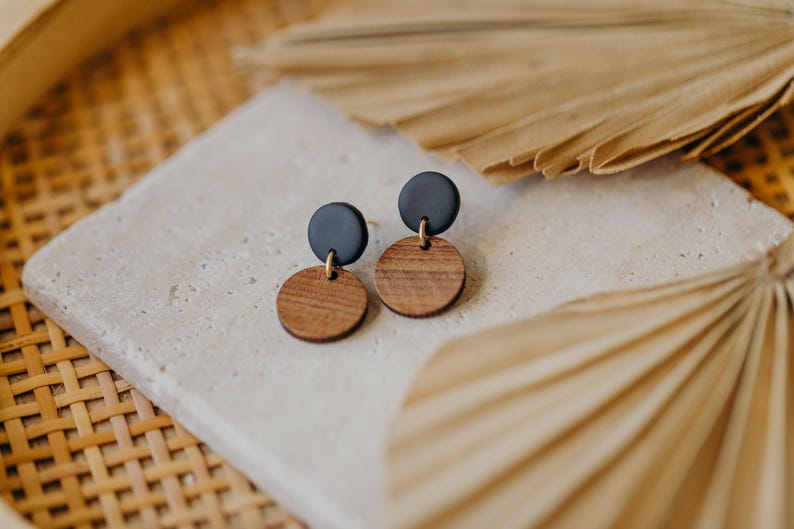 May include: A pair of earrings with black and brown wooden circles. The earrings are on a white surface with a brown woven background.