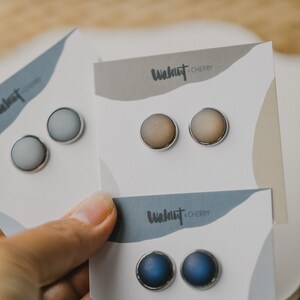 Op de afbeelding: Drie paar oorbel studs op witte kaartjes. De kaartjes hebben de tekst "Walnut & Cherry" erop gedrukt. De oorbel studs zijn in tinten blauw, grijs en bruin.