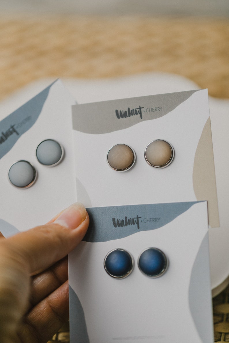 Op de afbeelding: Drie paar oorbel studs op witte kaartjes. De kaartjes hebben de tekst "Walnut & Cherry" erop gedrukt. De oorbel studs zijn in tinten blauw, grijs en bruin.