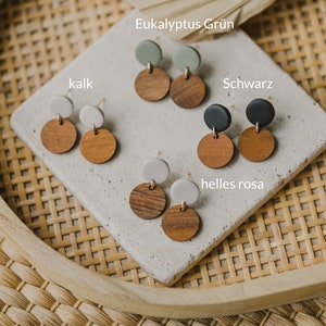 May include: Four pairs of earrings with wooden and clay circles. The colors are black, white, green, and light pink. The text "Eukalyptus Gr&uuml;n", "kalk", "Schwarz", "helles rosa" is visible.
