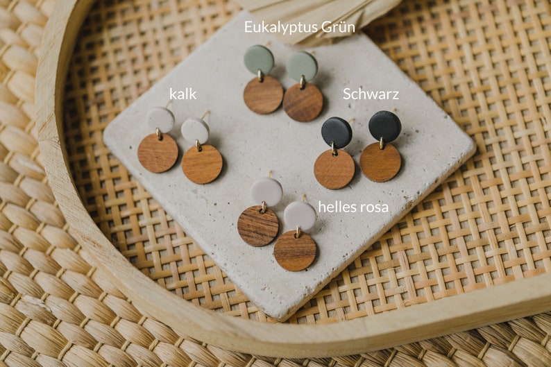 May include: Four pairs of earrings with wooden and clay circles. The colors are black, white, green, and light pink. The text "Eukalyptus Gr&uuml;n", "kalk", "Schwarz", "helles rosa" is visible.