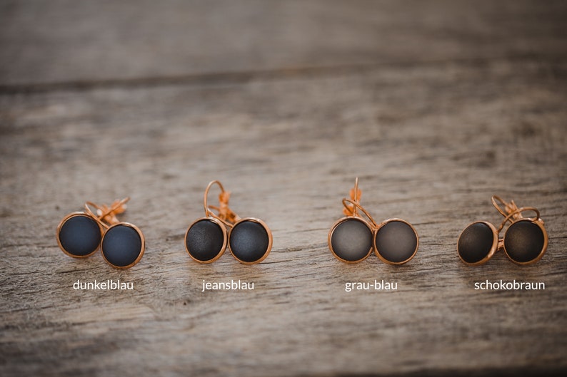 May include: Four pairs of earrings with different colored glass cabochons. The earrings are in shades of blue and brown. The colors are labeled "dunkelblau", "jeansblau", "grau-blau", and "schokobraun".