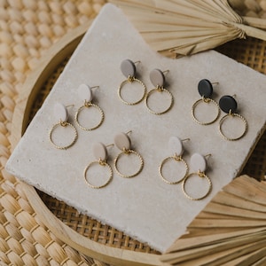 May include: Six pairs of gold hoop earrings with a twisted wire design. Each pair features a different colored round clay charm in beige, gray, black, and white.