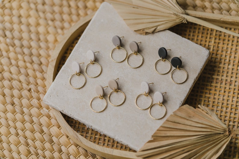 May include: Six pairs of gold hoop earrings with a twisted wire design. Each pair features a different colored round clay charm in beige, gray, black, and white.