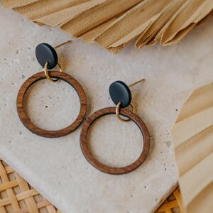 May include: A pair of wooden hoop earrings with a black disc accent and gold-colored hardware. The earrings are displayed on a light-colored stone surface, with dried palm leaves in the background. The earrings have a minimalist, natural aesthetic.
